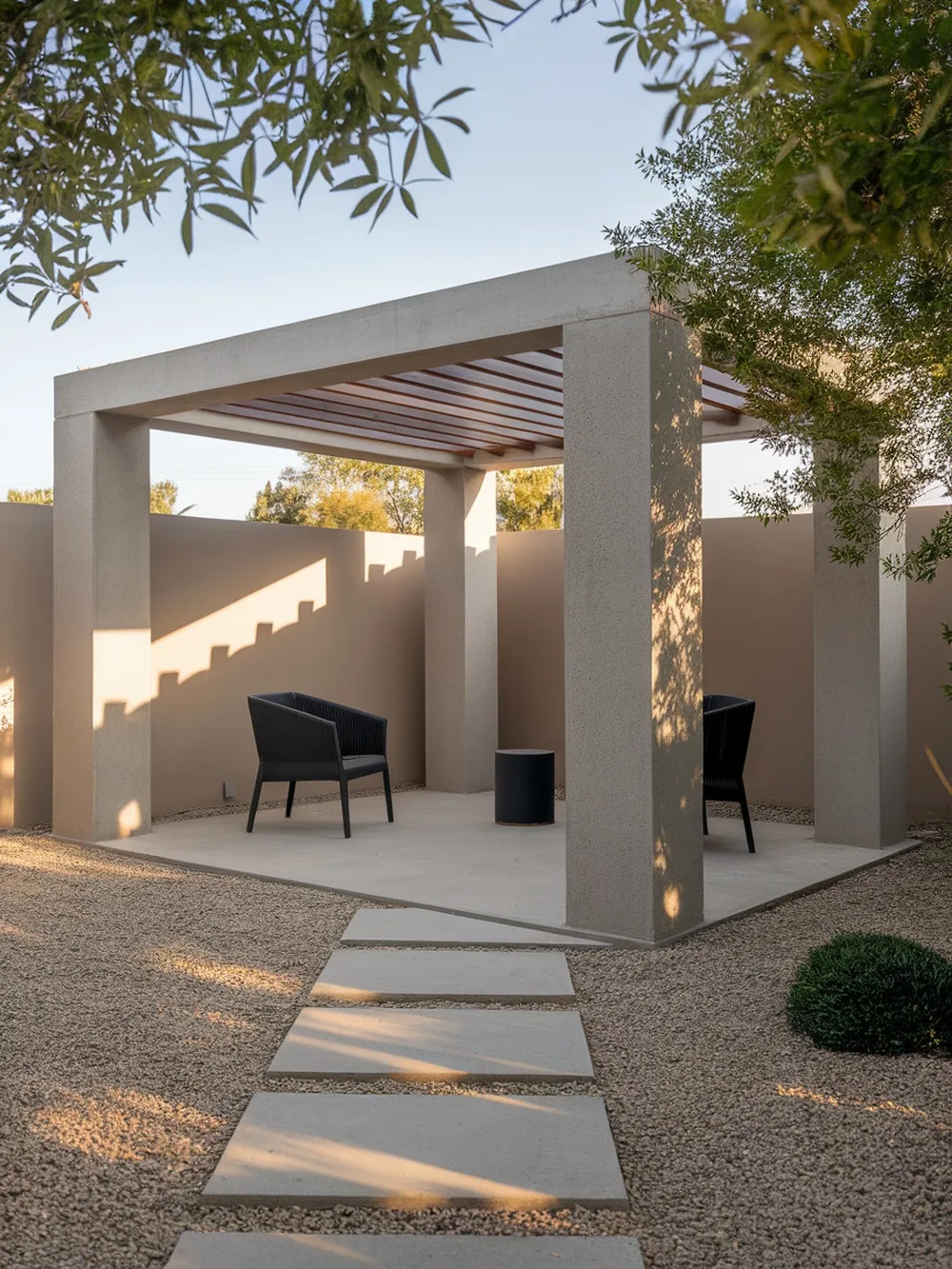 A minimalist pergola with concrete columns, featuring two black chairs and a small table, surrounded by gravel and greenery.