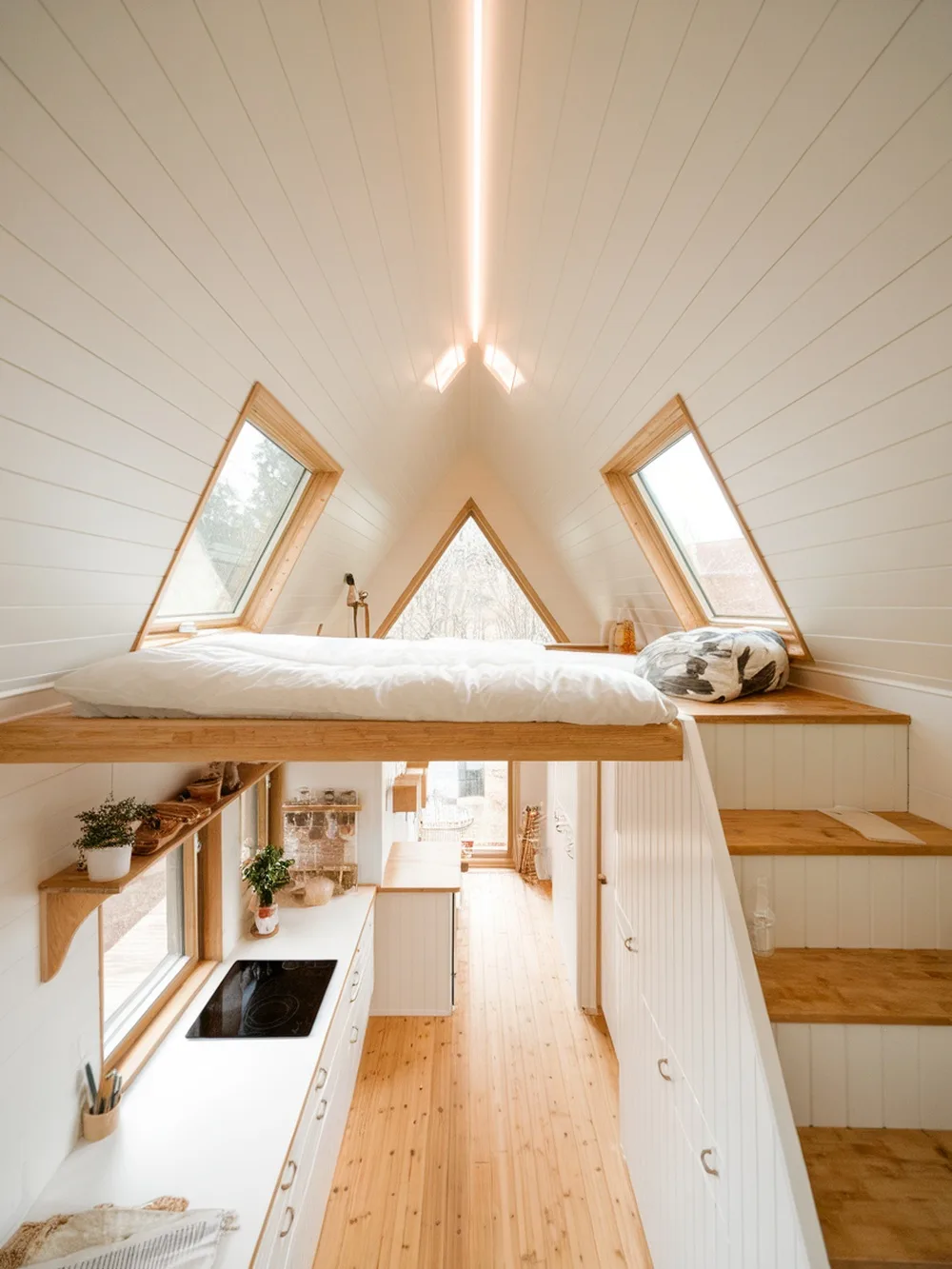 Interior of a minimalist tiny house featuring a lofted bed, large windows, and a compact kitchen.